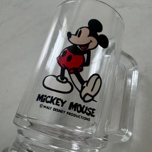 May include: A clear glass mug featuring a graphic of Mickey Mouse wearing red shorts. Below the character, the text reads MICKEY MOUSE and WALT DISNEY PRODUCTIONS. The mug has a sturdy handle and a smooth, transparent finish, showcasing the iconic character design.