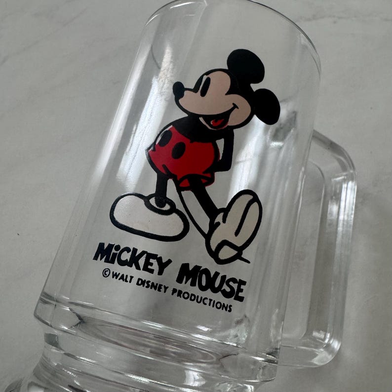 May include: A clear glass mug featuring a graphic of Mickey Mouse wearing red shorts. Below the character, the text reads MICKEY MOUSE and WALT DISNEY PRODUCTIONS. The mug has a sturdy handle and a smooth, transparent finish, showcasing the iconic character design.