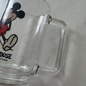May include: A clear glass mug featuring a graphic of Mickey Mouse in black, red, and white. The mug has a sturdy, rectangular handle and a base. The text "MOUSE" and "CTIONS" is printed in black on the side of the glass near the bottom.