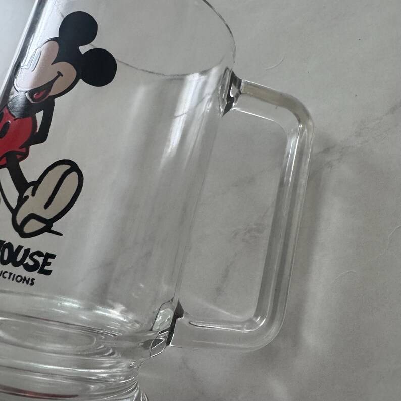 May include: A clear glass mug featuring a graphic of Mickey Mouse in black, red, and white. The mug has a sturdy, rectangular handle and a base. The text "MOUSE" and "CTIONS" is printed in black on the side of the glass near the bottom.