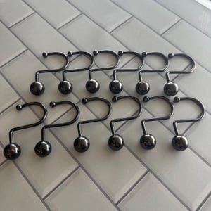 May include: A set of ten black metal shower curtain hooks. Each hook is shaped like a question mark, with a small ball at the bottom and a curved top for hanging. The hooks are arranged on a light gray tiled surface.