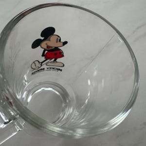 May include: A clear glass mug featuring a graphic of Mickey Mouse on the interior wall. The character is depicted in black, red, and tan colors. The text MICKEY MOUSE is printed in black below the character. The mug has a smooth, transparent finish.