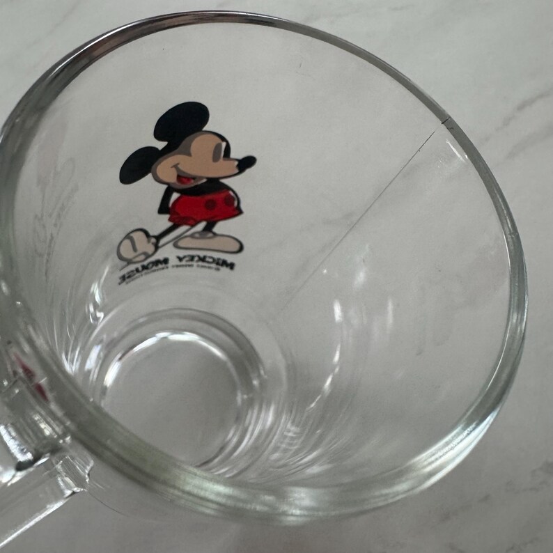 May include: A clear glass mug featuring a graphic of Mickey Mouse on the interior wall. The character is depicted in black, red, and tan colors. The text MICKEY MOUSE is printed in black below the character. The mug has a smooth, transparent finish.