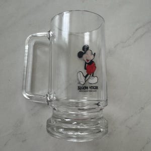 May include: A clear glass beer mug featuring a graphic of Mickey Mouse wearing red shorts and yellow shoes. The text MICKEY MOUSE is printed in black below the character, followed by smaller text reading WALT DISNEY PRODUCTIONS.