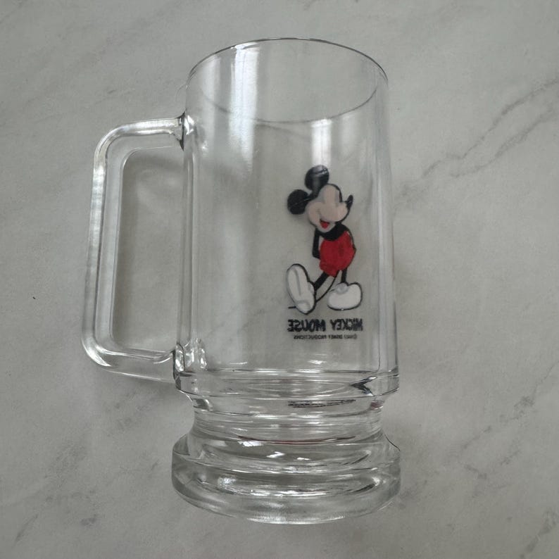 May include: A clear glass beer mug featuring a graphic of Mickey Mouse wearing red shorts and yellow shoes. The text MICKEY MOUSE is printed in black below the character, followed by smaller text reading WALT DISNEY PRODUCTIONS.