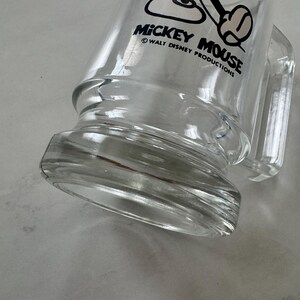 May include: A clear glass mug featuring a black graphic of Mickey Mouse and the text MICKEY MOUSE &copy; WALT DISNEY PRODUCTIONS printed on the side. The mug has a thick, sturdy base and a side handle, showcasing a transparent design throughout.