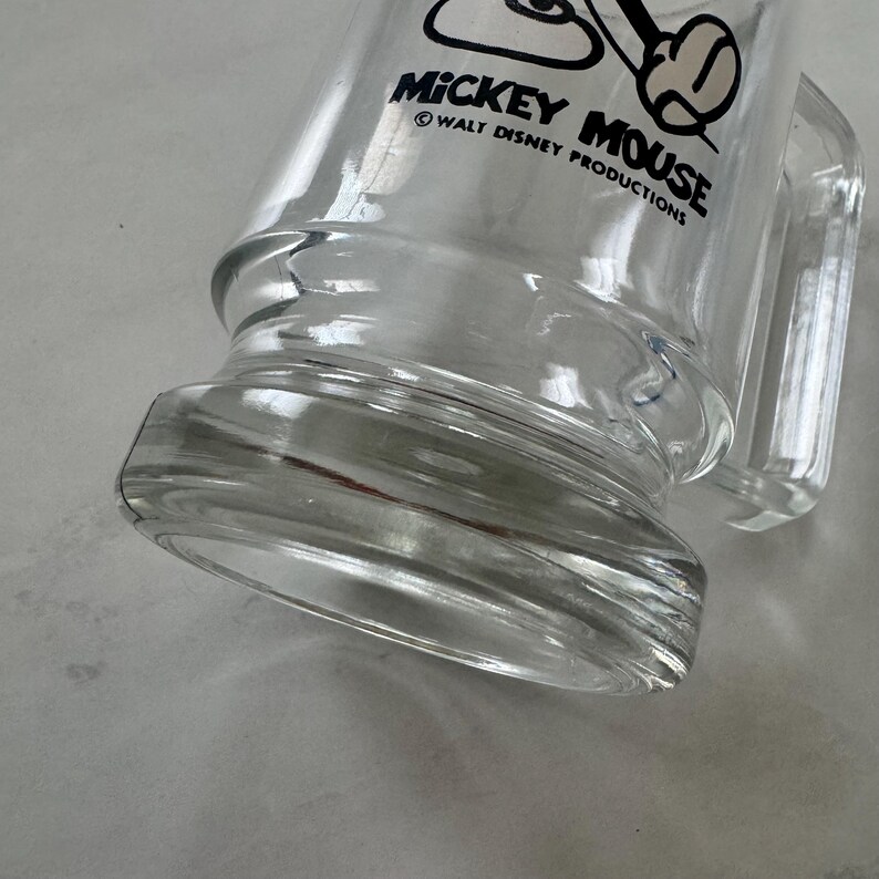 May include: A clear glass mug featuring a black graphic of Mickey Mouse and the text MICKEY MOUSE &copy; WALT DISNEY PRODUCTIONS printed on the side. The mug has a thick, sturdy base and a side handle, showcasing a transparent design throughout.
