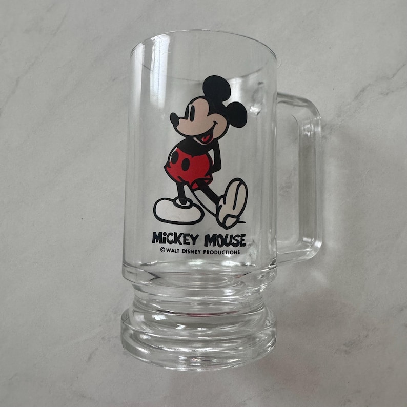 May include: A clear glass beer mug featuring a graphic of Mickey Mouse in red shorts and yellow shoes. The text MICKEY MOUSE and WALT DISNEY PRODUCTIONS is printed in black below the character. The mug has a sturdy handle and a thick base.