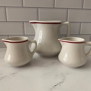 May include: Three white ceramic pitchers with a thin red band around the top. The pitchers are of different sizes, with the largest pitcher being a traditional milk pitcher shape.