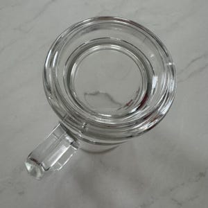 May include: A clear glass beer mug viewed from directly above, featuring a thick circular rim, a smooth interior base, and a sturdy rectangular handle extending from the side. The transparent glass construction shows a simple, functional design.