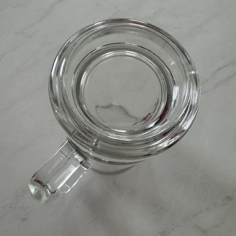May include: A clear glass beer mug viewed from directly above, featuring a thick circular rim, a smooth interior base, and a sturdy rectangular handle extending from the side. The transparent glass construction shows a simple, functional design.
