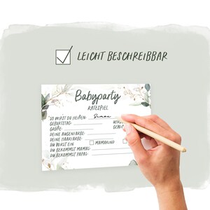 May include: A white card with a green and gold floral design. The card has the text "Babyparty Ratespiel" and a list of questions to guess about a baby, such as "So wirst du hei&szlig;en", "Geburtstag", "Gr&ouml;&szlig;e", "Deine Augenfarbe", "Deine Haarfarbe", "Du wirst ein", "Du bekommst Mamas", and "Du bekommst Papas".
