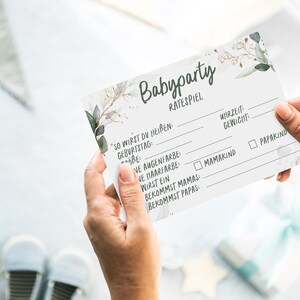 May include: A white card with a floral design and the text "Babyparty Ratespiel" printed on it. The card has a list of questions to guess about a baby, such as the baby's name, birthdate, eye color, hair color, and gender.