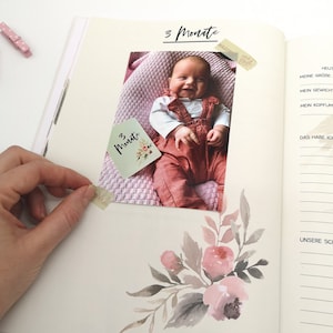 May include: A baby girl wearing a red overall and white shirt is lying on a pink blanket. The photo is in a scrapbook with a handwritten note that says "3 Monate" which is German for "3 months".