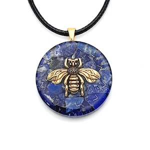 May include: A gold bee pendant with a blue lapis lazuli stone background. The pendant is on a black cord.