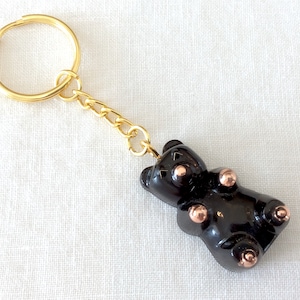 Könnte beinhalten: Schwarzer Gummibärchen-Schlüsselanhänger mit goldener Kette und Ring. Der Gummibär hat eine glänzende schwarze Oberfläche und kupferfarbene Akzente.