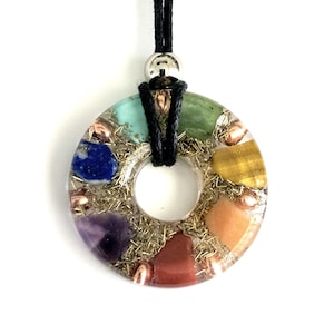 May include: A round, clear resin pendant with seven chakra stones embedded in it. The stones are arranged in a circle and are surrounded by gold glitter. The pendant is attached to a black cord.