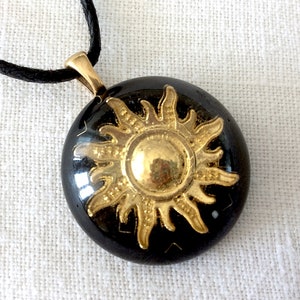 May include: A round, black pendant with a gold-colored sun design. The pendant hangs from a black cord. The sun has a detailed, radiant design. The pendant is likely made of resin or a similar material.