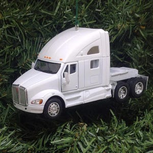 May include: A white semi-truck cab ornament featuring a detailed silver grille, orange marker lights, and black wheels with silver rims. The vehicle model includes a sleeper cab section, side mirrors, and a green string attached for hanging.