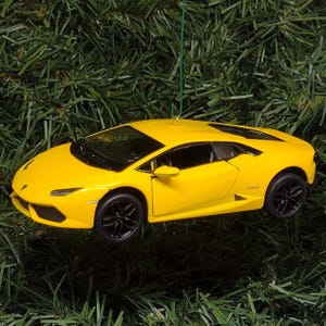May include: A bright yellow sports car model ornament featuring a sleek aerodynamic body, black multi-spoke wheels, and a low-profile design. The miniature vehicle is suspended by a thin green string, showcasing sharp angular headlights and side vents.