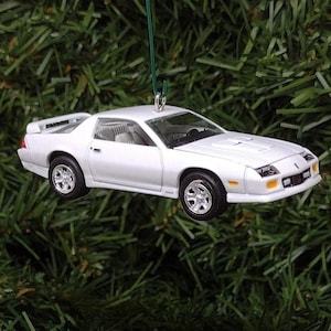 May include: A white model car ornament featuring a sleek coupe design with a rear spoiler, silver-toned wheels, and yellow side marker lights. The miniature vehicle is suspended by a thin green string, showcasing a detailed exterior and interior.