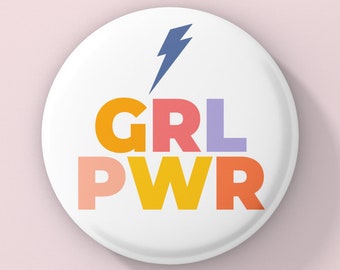Puede incluir: Un botón blanco con un rayo azul y el texto "GRL PWR" en letras de colores. Las letras están apiladas una encima de la otra, con "GRL" en naranja, rosa y morado, y "PWR" en amarillo, rosa y rojo.
