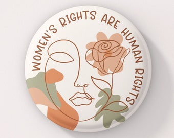 Puede incluir: Un botón blanco con un dibujo de línea marrón de una cara y una rosa. El texto "Women's Rights Are Human Rights" está escrito alrededor del borde del botón.