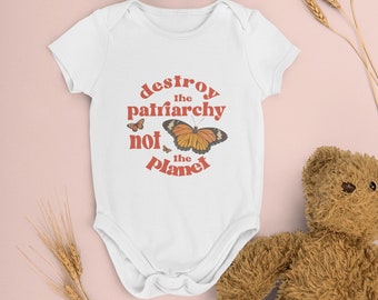 May include: White baby onesie with a red graphic that says "destroy the patriarchy not the planet" with a butterfly illustration. The onesie is on a pink background with a brown teddy bear and wheat stalks.