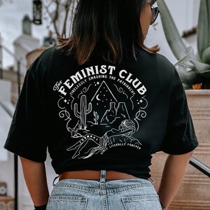 May include: Black t-shirt with a white graphic design featuring a desert scene with the text "The Feminist Club Tirelessly Smashing The Patriarchy Since Literally Forever".