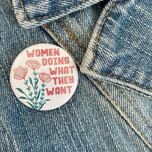 May include: A white button with a pink floral design and the text "Women Doing What They Want" in red lettering.