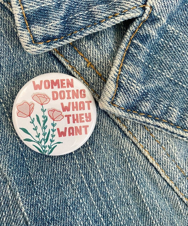 May include: A white button with a pink floral design and the text "Women Doing What They Want" in red lettering.