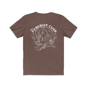 May include: A brown t-shirt with a white graphic design. The graphic features a desert scene with a cactus, mountains, and a crescent moon. The text "The Feminist Club" is written in a circular design around the scene. The text "Tirelessly Smashing the Patriarchy" is written in a smaller font below the scene. The text "Once Literally Forever" is written in a smaller font below the scene.