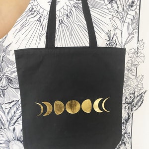 May include: Black canvas tote bag with gold moon phases printed on the front. The bag has black straps and the text "THE SUN." is printed on the bottom.