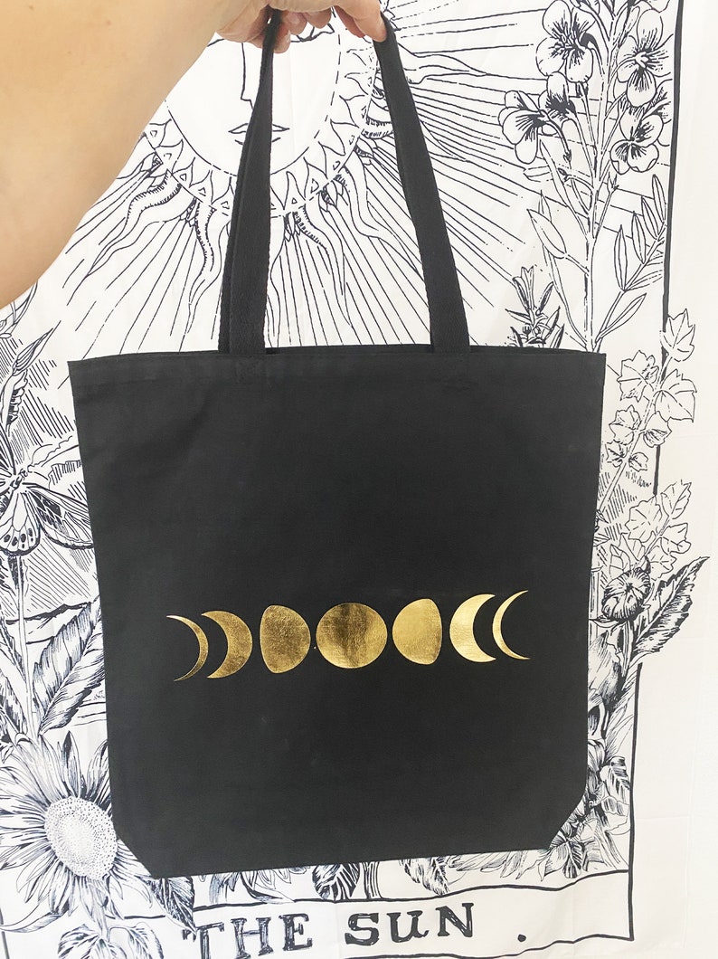 May include: Black canvas tote bag with gold moon phases printed on the front. The bag has black straps and the text "THE SUN." is printed on the bottom.