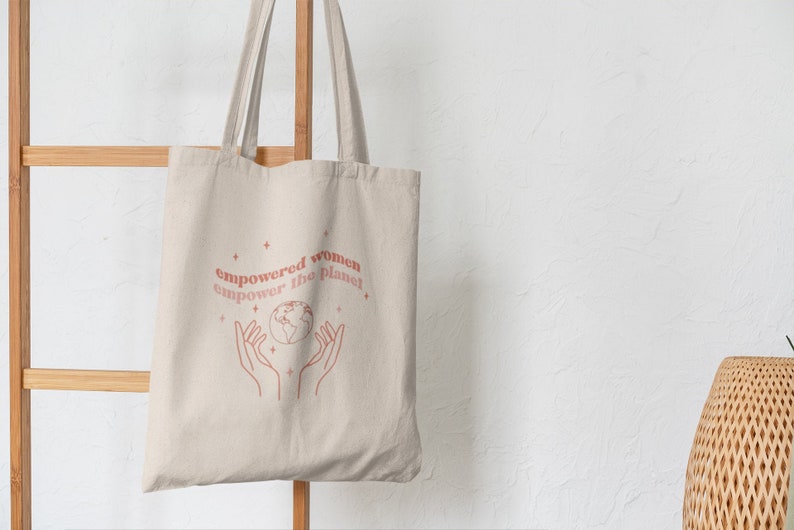 Może przedstawiać: Beżowa torba na zakupy z napisem "empowered women empower the planet" i grafiką dw&oacute;ch dłoni trzymających globus.
