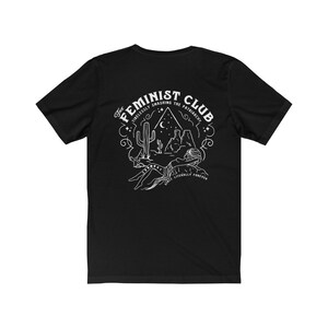 May include: Black t-shirt featuring a white graphic design of a desert scene with a cactus, mountains, and a crescent moon. The text "The Feminist Club" is in a circular design around the scene. The text "Tirelessly Smashing the Patriarchy" is in a smaller font above the scene. The text "Once Literally Forever" is in a smaller font below the scene.