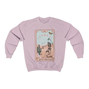 Pode incluir: Uma camisa de frio rosa claro com um design de carta de tar&ocirc; que apresenta um esqueleto dan&ccedil;ando em uma paisagem des&eacute;rtica. O cart&atilde;o diz "IV Morte ao Patriarcado".