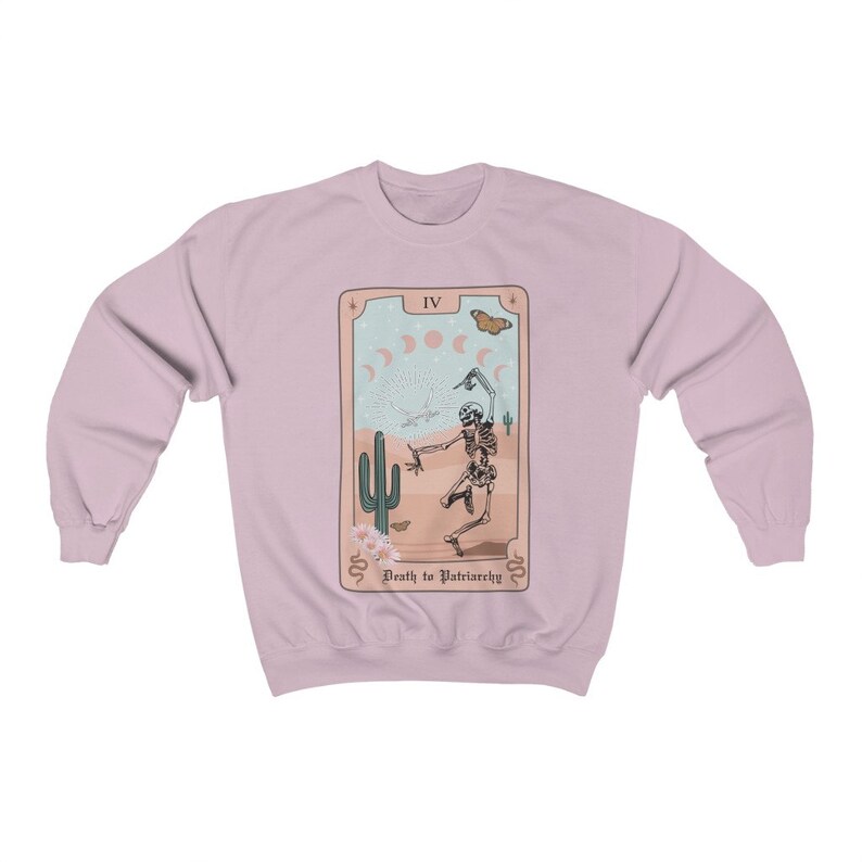 Pode incluir: Uma camisa de frio rosa claro com um design de carta de tar&ocirc; que apresenta um esqueleto dan&ccedil;ando em uma paisagem des&eacute;rtica. O cart&atilde;o diz "IV Morte ao Patriarcado".