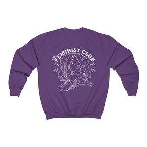 Może przedstawiać: Fioletowa bluza z białym nadrukiem graficznym, na kt&oacute;rym widnieje napis "The Feminist Club" wraz ze sceną pustynną i tekstem "Tirelessly Smashing the Patriarchy Since Literally Forever".