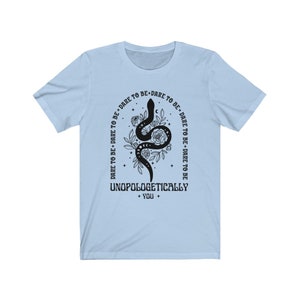 Pu&ograve; includere: Maglietta blu chiaro con un serpente nero e un disegno floreale. Il testo "DARE TO BE+DARE TO BE DARE TO BE+DARE TO BE TO BE DARE TO BE UNOPOLOGETICALLY + YOU +" &egrave; stampato sulla maglietta.
