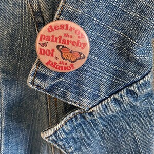 May include: A pink button with the text "Destroy the patriarchy not the planet" in red and white lettering. A butterfly with orange and black wings is in the center of the button.