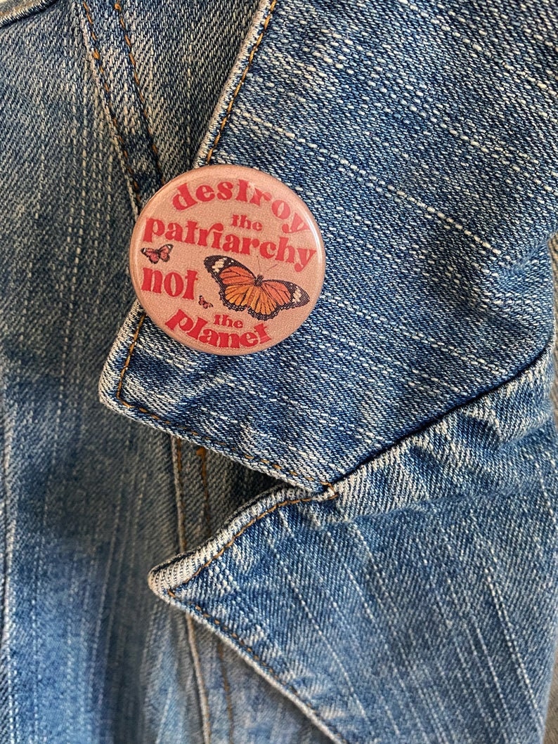 May include: A pink button with the text "Destroy the patriarchy not the planet" in red and white lettering. A butterfly with orange and black wings is in the center of the button.