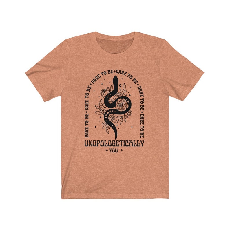 Pu&ograve; includere: Una maglietta marrone chiaro con un serpente nero e un disegno floreale. Il testo "DARE TO BE+DARE TO BE+DARE TO BE+DARE TO BE+DARE TO BE" &egrave; stampato in un cerchio attorno al serpente. Il testo "UNOPOLOGETICALLY + YOU +" &egrave; stampato sotto il serpente.
