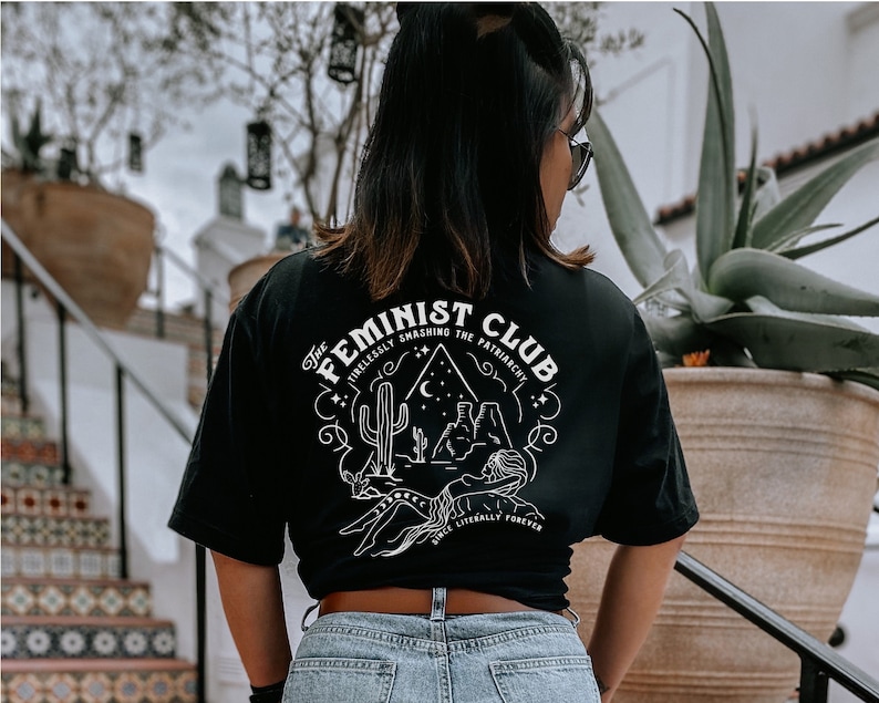 May include: Black t-shirt with a white graphic design featuring a desert scene with the text "The Feminist Club Tirelessly Smashing The Patriarchy Since Literally Forever".