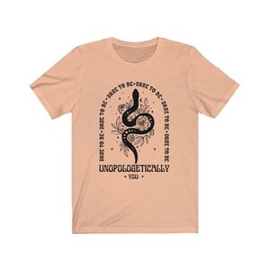 Pu&ograve; includere: T-shirt color pesca con un designo di serpente nero e il testo "DARE TO BE+DARE TO BE+DARE TO BE+DARE TO BE" che circonda il serpente. Il testo "UNOPOLOGETICALLY + YOU +" &egrave; sotto il serpente.