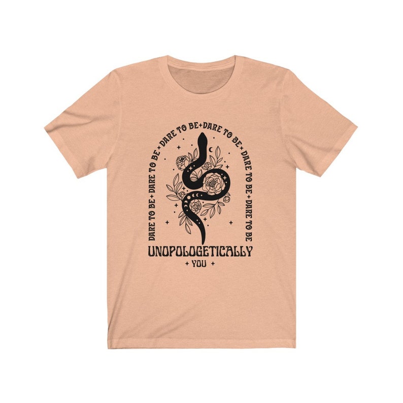 Pu&ograve; includere: T-shirt color pesca con un designo di serpente nero e il testo "DARE TO BE+DARE TO BE+DARE TO BE+DARE TO BE" che circonda il serpente. Il testo "UNOPOLOGETICALLY + YOU +" &egrave; sotto il serpente.
