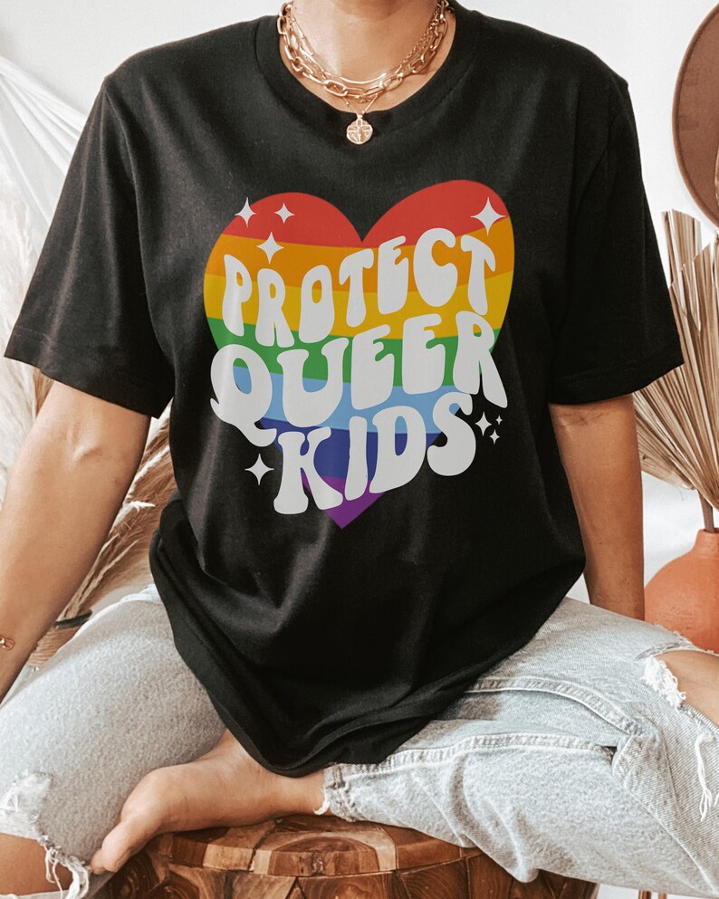 Puede incluir: Camiseta negra con un gr&aacute;fico de coraz&oacute;n arco&iacute;ris que dice "Proteger a los ni&ntilde;os queer".