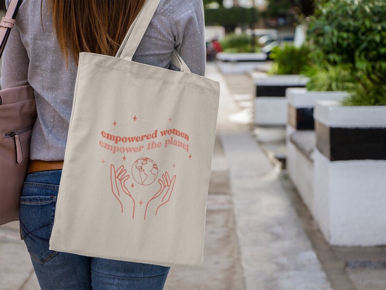 Może przedstawiać: Biała torba typu tote z r&oacute;żowym wzorem, na kt&oacute;rym widnieje napis "empowered women empower the planet" oraz stylizowany obraz dłoni trzymających globus.