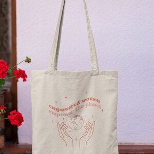 Może przedstawiać: Beżowa torba typu tote z r&oacute;żowym wzorem z tekstem "empowered women empower the planet" i stylizowanym obrazem dłoni trzymających globus.