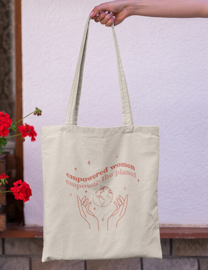 Może przedstawiać: Beżowa torba typu tote z r&oacute;żowym wzorem z tekstem "empowered women empower the planet" i stylizowanym obrazem dłoni trzymających globus.